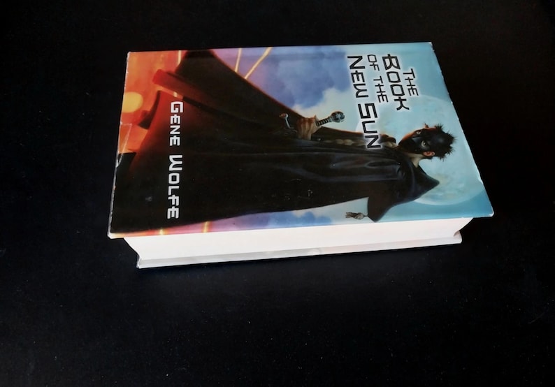 Puede incluir: Un libro de tapa dura titulado "The Book of the New Sun" de Gene Wolfe. La portada presenta una figura con una capa oscura y una espada, sobre un cielo azul y naranja. El libro est&aacute; sobre una superficie negra.