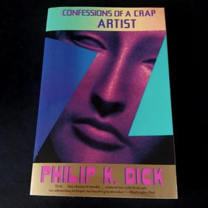 Peut inclure: Un livre de poche intitulé "CONFESSIONS OF A CRAP ARTIST" de Philip K. Dick. La couverture présente un visage stylisé dans des tons violets et roses, avec des formes géométriques bleues et turquoise. Le nom de l'auteur est en rose.