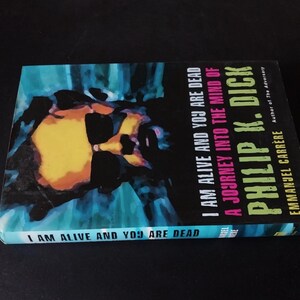 以下が含まれることがあります： フィリップ・K・ディック著「I Am Alive and You Are Dead」というタイトルの本。表紙は青、黄、黒色を使った抽象的なデザイン。著者はエマニュエル・カレレ。タイトルは背表紙にも記載。