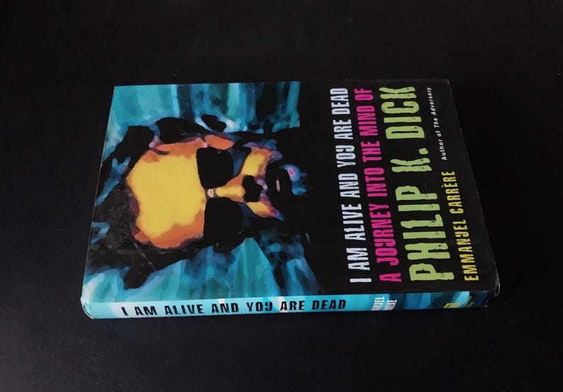 以下が含まれることがあります： フィリップ・K・ディック著「I Am Alive and You Are Dead」というタイトルの本。表紙は青、黄、黒色を使った抽象的なデザイン。著者はエマニュエル・カレレ。タイトルは背表紙にも記載。