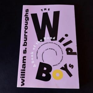 May include: A paperback book titled "The Wild Boys" by William S. Burroughs. The cover is light purple with large black letters spelling out "WILD BOYS". The title is in a bold, sans-serif font. The author's name is printed vertically on the left side.