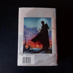 Puede incluir: Contraportada de un libro con una ilustraci&oacute;n de fantas&iacute;a. Una figura con una capa oscura sostiene una espada contra un cielo iluminado por la luna y una puesta de sol ardiente. El ISBN y el c&oacute;digo de barras son visibles.