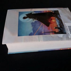 Puede incluir: Un libro de tapa dura con una sobrecubierta que presenta una ilustraci&oacute;n de fantas&iacute;a. La portada muestra una silueta sobre un fondo de luna, nubes y un cielo colorido. El libro est&aacute; sobre una superficie negra.