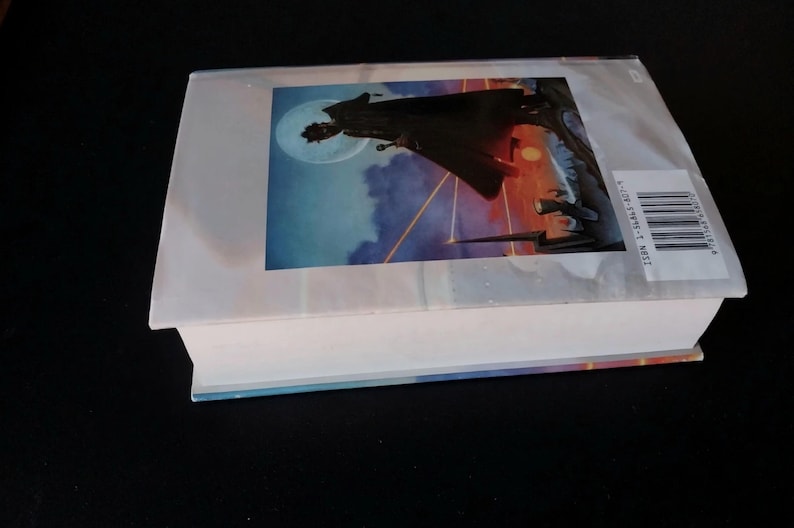 Puede incluir: Un libro de tapa dura con una sobrecubierta que presenta una ilustraci&oacute;n de fantas&iacute;a. La portada muestra una silueta sobre un fondo de luna, nubes y un cielo colorido. El libro est&aacute; sobre una superficie negra.