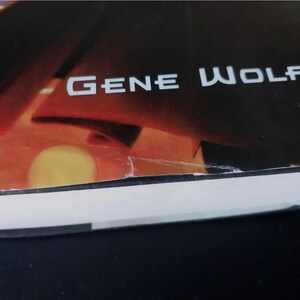 Puede incluir: Una portada de libro con el nombre del autor, "GENE WOLFE", en letras blancas. El fondo presenta un degradado de naranja, rojo y negro, con una forma amarilla circular. Las p&aacute;ginas del libro son visibles en la parte inferior.