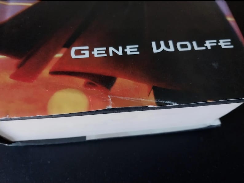 Puede incluir: Una portada de libro con el nombre del autor, "GENE WOLFE", en letras blancas. El fondo presenta un degradado de naranja, rojo y negro, con una forma amarilla circular. Las p&aacute;ginas del libro son visibles en la parte inferior.