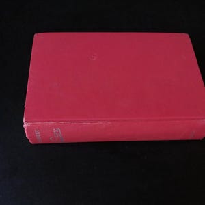 以下が含まれることがあります： 鮮やかな赤色の表紙の、閉じられた長方形の本。本の背表紙には、著者の名前とタイトルが銀色の文字で表示されています。本は黒い背景に置かれ、その色と形が際立っています。