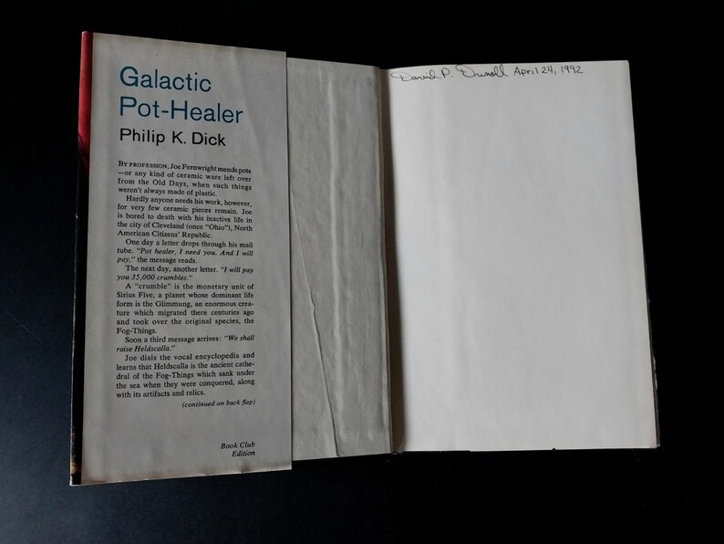 以下が含まれることがあります： フィリップ・K・ディック著「Galactic Pot-Healer」というタイトルの開かれた本。表紙は明るい色で黒い文字が書かれています。この本はブッククラブ版です。内側のページには手書きで「Donald P. Chinell April 24, 1992」と書かれています。