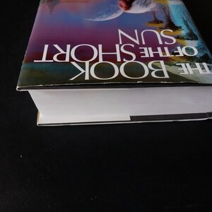 Puede incluir: Se muestra un libro de tapa dura titulado "THE BOOK OF THE SHORT SUN". La portada presenta una ilustraci&oacute;n colorida con el t&iacute;tulo en letras blancas. Las p&aacute;ginas del libro son blancas y el lomo es visible.