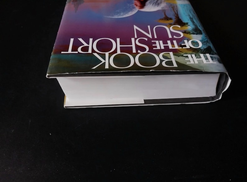 Puede incluir: Se muestra un libro de tapa dura titulado "THE BOOK OF THE SHORT SUN". La portada presenta una ilustraci&oacute;n colorida con el t&iacute;tulo en letras blancas. Las p&aacute;ginas del libro son blancas y el lomo es visible.
