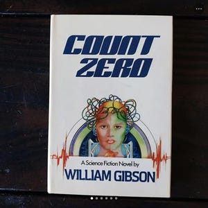 Puede incluir: Una cubierta de libro blanca con el título "COUNT ZERO" en letras azules. La cubierta presenta una ilustración colorida de una cara con una corona de cables de color arcoíris. El texto "A Science Fiction Novel by WILLIAM GIBSON" está impreso debajo de la ilustración.