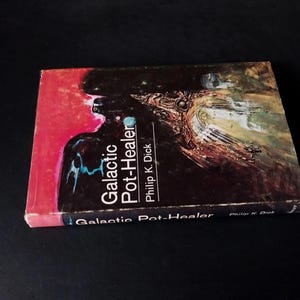 以下が含まれることがあります： フィリップ・K・ディック著の「Galactic Pot-Healer」というタイトルの本。表紙は赤と黒の抽象的なデザインで、タイトルは白と青で書かれています。本は暗い表面にあります。