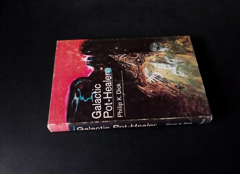 以下が含まれることがあります： フィリップ・K・ディック著の「Galactic Pot-Healer」というタイトルの本。表紙は赤と黒の抽象的なデザインで、タイトルは白と青で書かれています。本は暗い表面にあります。