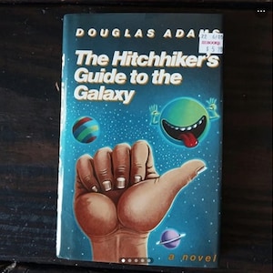 May include: A blue paperback book with a white title, "The Hitchhiker's Guide to the Galaxy" by Douglas Adams. The cover features a cartoon hand with a planet in the background.