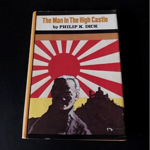 Peut inclure: Un livre de poche vintage intitulé "The Man In The High Castle" de Philip K. Dick. La couverture présente un portrait d'homme, un motif de soleil levant et une silhouette de château. Le livre a un dos jaune et est posé sur un fond noir.