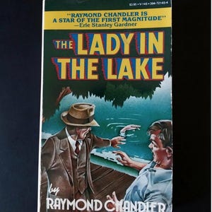 Può includere: Copertina di un libro tascabile vintage per "The Lady in the Lake" di Raymond Chandler. La copertina presenta un uomo in giacca e cappello, un altro uomo e una scena lacustre. Il titolo è in grandi lettere colorate.