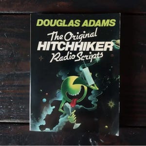 Könnte beinhalten: Ein schwarzer und grüner Buchumschlag mit dem Titel "The Original Hitchhiker Radio Scripts" von Douglas Adams. Der Umschlag zeigt eine Comicfigur mit einem grünen Kopf und einer roten Zunge.
