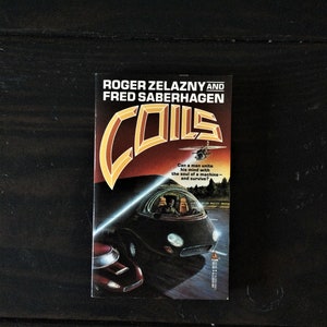 May include: A paperback book cover with a black and orange design. The title of the book is "Coils" by Roger Zelazny and Fred Saberhagen. The cover features a futuristic car driving on a road with a bright light shining from the front of the car.