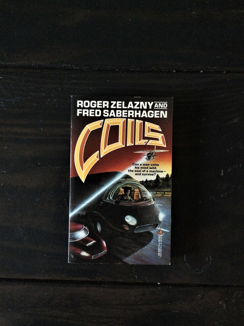 May include: A paperback book cover with a black and orange design. The title of the book is "Coils" by Roger Zelazny and Fred Saberhagen. The cover features a futuristic car driving on a road with a bright light shining from the front of the car.