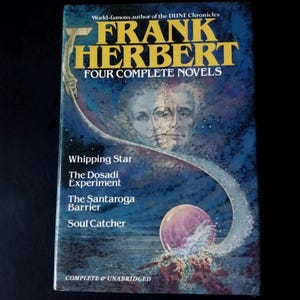 Puede incluir: Un libro de bolsillo titulado "FRANK HERBERT FOUR COMPLETE NOVELS" con una cubierta azul y blanca. La cubierta presenta el nombre del autor en letras amarillas grandes, un retrato y los títulos de cuatro novelas: Whipping Star, The Dosadi Experiment, The Santaroga Barrier y Soul Catcher.