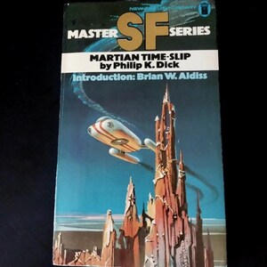 Peut inclure: Un livre de poche vintage intitulé "Martian Time-Slip" de Philip K. Dick. La couverture présente une illustration de science-fiction rétro d'un vaisseau spatial et d'une structure imposante sur fond bleu. Fait partie de la "SF Series".