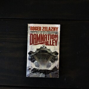 May include: A paperback book cover with a red and white title "Damnation Alley" by Roger Zelazny, the Hugo Nebula-winning author of Lord of Light and the Chronicles of Amber. The cover features a futuristic vehicle driving through a desolate landscape.