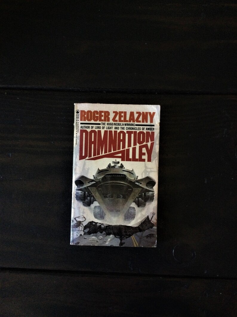 May include: A paperback book cover with a red and white title "Damnation Alley" by Roger Zelazny, the Hugo Nebula-winning author of Lord of Light and the Chronicles of Amber. The cover features a futuristic vehicle driving through a desolate landscape.