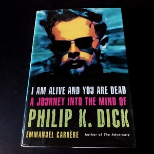 以下が含まれることがあります： サングラスをかけた男性の肖像画が描かれた本の表紙。「I AM ALIVE AND YOU ARE DEAD A JOURNEY INTO THE MIND OF PHILIP K. DICK」エマニュエル・カレールの著書。