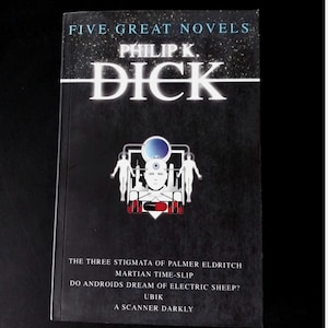 Puede incluir: Un libro de tapa dura negro titulado "FIVE GREAT NOVELS" de Philip K. Dick. La portada presenta texto blanco y un gráfico de una figura robótica. Los títulos de las novelas incluyen "Ubik" y "A Scanner Darkly".