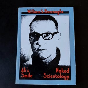 Pode incluir: Um livro de bolso com uma borda azul clara apresenta um retrato a preto e branco de um homem com óculos. O título "William S. Burroughs" está no topo, com "Ali's Smile" e "Naked Scientology" abaixo do retrato.