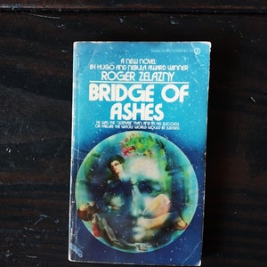 May include: A blue and white paperback book cover with the title "Bridge of Ashes" by Roger Zelazny. The cover features a colorful illustration of a face surrounded by a blue sphere.