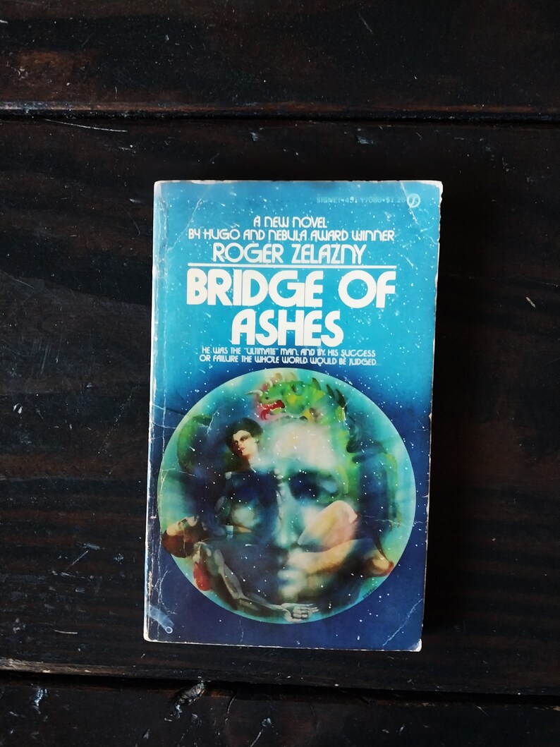 May include: A blue and white paperback book cover with the title "Bridge of Ashes" by Roger Zelazny. The cover features a colorful illustration of a face surrounded by a blue sphere.