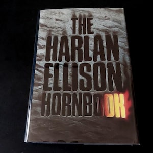 May include: A book titled "THE HARLAN ELLISON HORNBOK" with a dark cover. The title is in large, bold, brown letters, set against a textured, grey background. The word "HORNBOK" is in a yellow and orange gradient.