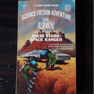 Op de afbeelding: Een vintage paperback boekomslag met een sciencefiction-avontuurroman met de titel "David Starr-Space Ranger" van Isaac Asimov, die schrijft als Paul French. De omslagillustratie toont een futuristisch voertuig en twee astronauten in groene jumpsuits en blauwe helmen.