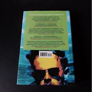以下が含まれることがあります： エマニュエル・カレールの著書「The Adversary」の裏表紙。サングラスをかけた男性の肖像画が、緑と青の背景に描かれています。本のレビューや引用文も含まれています。