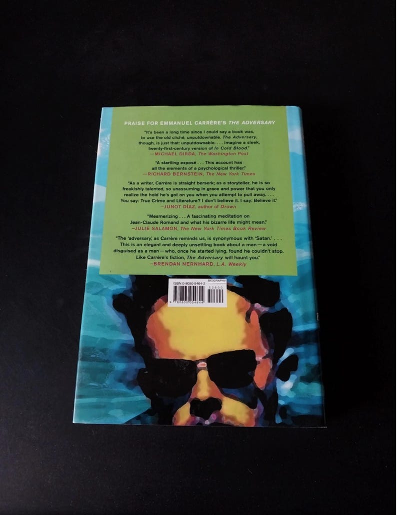 以下が含まれることがあります： エマニュエル・カレールの著書「The Adversary」の裏表紙。サングラスをかけた男性の肖像画が、緑と青の背景に描かれています。本のレビューや引用文も含まれています。