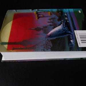 Puede incluir: Un libro de tapa dura con una ilustraci&oacute;n vibrante en la portada. La portada presenta un fondo rojo y amarillo con un paisaje urbano silueteado y un gran tibur&oacute;n estilizado. El lomo del libro incluye un c&oacute;digo de barras y un ISBN.