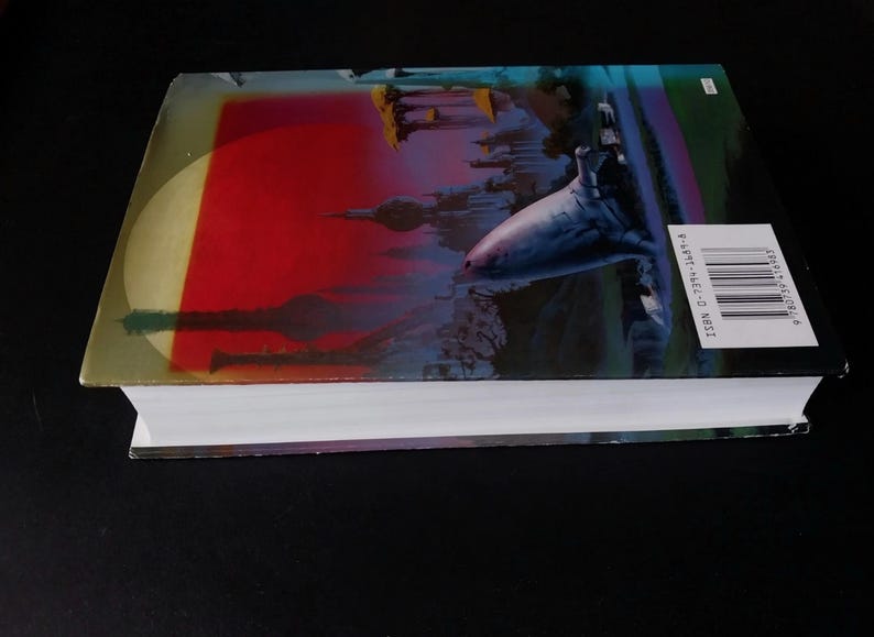 Puede incluir: Un libro de tapa dura con una ilustraci&oacute;n vibrante en la portada. La portada presenta un fondo rojo y amarillo con un paisaje urbano silueteado y un gran tibur&oacute;n estilizado. El lomo del libro incluye un c&oacute;digo de barras y un ISBN.