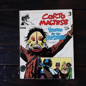 May include: A comic book cover featuring a colorful illustration of a masked figure with a menacing expression. The title of the comic is "Corto Maltese: Voodoo for the President" with the author's name "Hugo Pratt" in smaller font. The cover also includes the text "With an introduction by Frank Miller".
