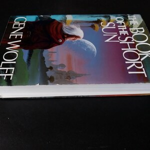 Puede incluir: Un libro de tapa dura titulado "The Book of the Short Sun" de Gene Wolfe. La portada presenta a una persona con cabello blanco y una capa roja, con un fondo celestial de una luna y un paisaje de fantas&iacute;a.