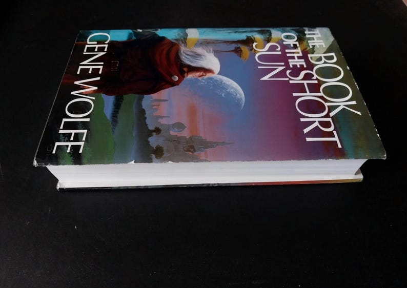Puede incluir: Un libro de tapa dura titulado "The Book of the Short Sun" de Gene Wolfe. La portada presenta a una persona con cabello blanco y una capa roja, con un fondo celestial de una luna y un paisaje de fantas&iacute;a.