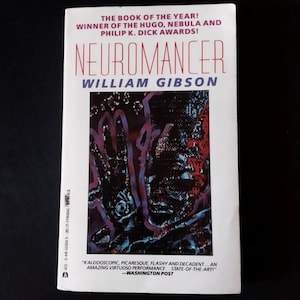 May include: A paperback book titled "Neuromancer" by William Gibson. The cover features a dark, abstract illustration with the title in large, red letters. The book is labeled "The Book of the Year!" and lists awards.