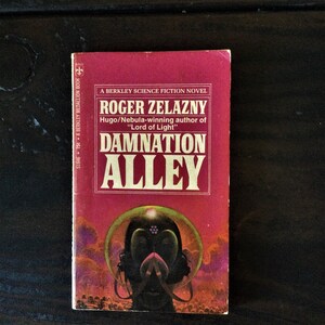 May include: A red paperback book with the title "Damnation Alley" by Roger Zelazny. The book cover features a futuristic illustration of a person wearing a helmet and a dark background.