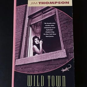 May include: A paperback book cover with a dark purple background featuring a woman looking out a window. The title "WILD TOWN" is written in gold letters. The author's name "JIM THOMPSON" is written in gold letters at the top of the cover.