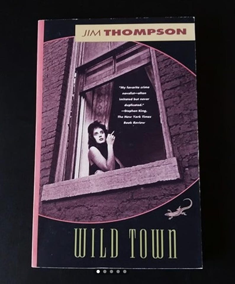 May include: A paperback book cover with a dark purple background featuring a woman looking out a window. The title "WILD TOWN" is written in gold letters. The author's name "JIM THOMPSON" is written in gold letters at the top of the cover.