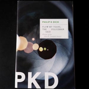 May include: A paperback book titled "Flow My Tears, The Policeman Said" by Philip K. Dick. The cover features a blurred, abstract design with a large black circle and the letters "PKD" in white.