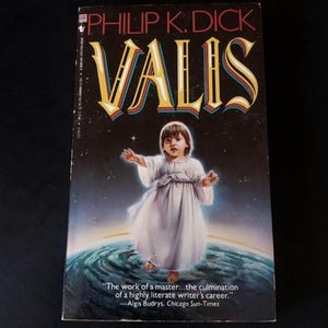 Peut inclure: Un livre de poche intitulé "VALIS" de Philip K. Dick. La couverture présente un enfant en robe blanche debout sur une planète, avec un fond étoilé. Le titre est en grandes lettres dorées stylisées. Le livre contient une citation d'Algis Budrys.