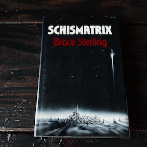 Puede incluir: Un libro de bolsillo negro con el título "Schismatrix" en blanco y el autor "Bruce Sterling" en rojo. La portada presenta una ilustración en blanco y negro de una ciudad futurista.