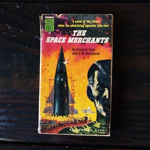 May include: A vintage paperback book cover with a rocket ship and a man's face. The book is titled "The Space Merchants" and is a novel of the future when advertising agencies take over. The cover is a mix of orange, yellow, red, and black.