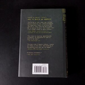 以下が含まれることがあります： David F. Dufty著「How to Build an Android」というタイトルの本の裏表紙。暗い色の表紙には、引用文やバーコードを含む白い文字が書かれています。この本は科学とロボット工学をテーマにしています。